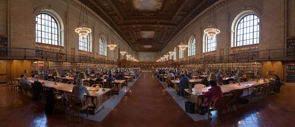 Readers study quietly at long wooden tables inside a bright public library reading room.